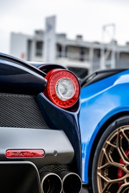 A close-up of a luxury sports car featuring a red tail light, dual exhaust pipes, and a portion of another car with a bright blue exterior and bronze wheels in the background. The focus is on the design elements and color contrast between the dark and vibrant hues.