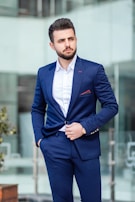 A well-dressed man in a blue suit and white shirt is standing outdoors, looking to the side. His hand is in his pocket, and he exudes confidence and style. The background is a blurred modern glass building.