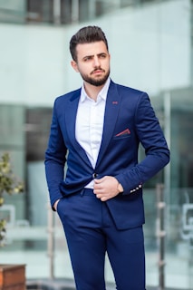A well-dressed man in a blue suit and white shirt is standing outdoors, looking to the side. His hand is in his pocket, and he exudes confidence and style. The background is a blurred modern glass building.