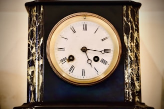 An antique clock with a white face and Roman numerals is set in a dark, ornate case with gold and marble-like detailing. The clock hands are simple and black, and the rim around the clock face is a polished bronze or gold color.