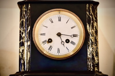 An antique clock with a white face and Roman numerals is set in a dark, ornate case with gold and marble-like detailing. The clock hands are simple and black, and the rim around the clock face is a polished bronze or gold color.