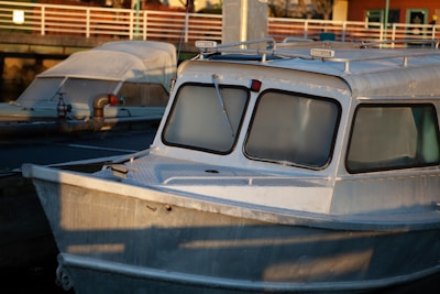 A docked metal boat with large frosted windows, slightly obscuring the inside of the cabin. The boat has a robust and functional design with an antenna and searchlights on the roof. Behind it, there is another boat with a more classic shape, covered with a white protective sheet. The background shows a wooden pier with a white railing and some buildings.