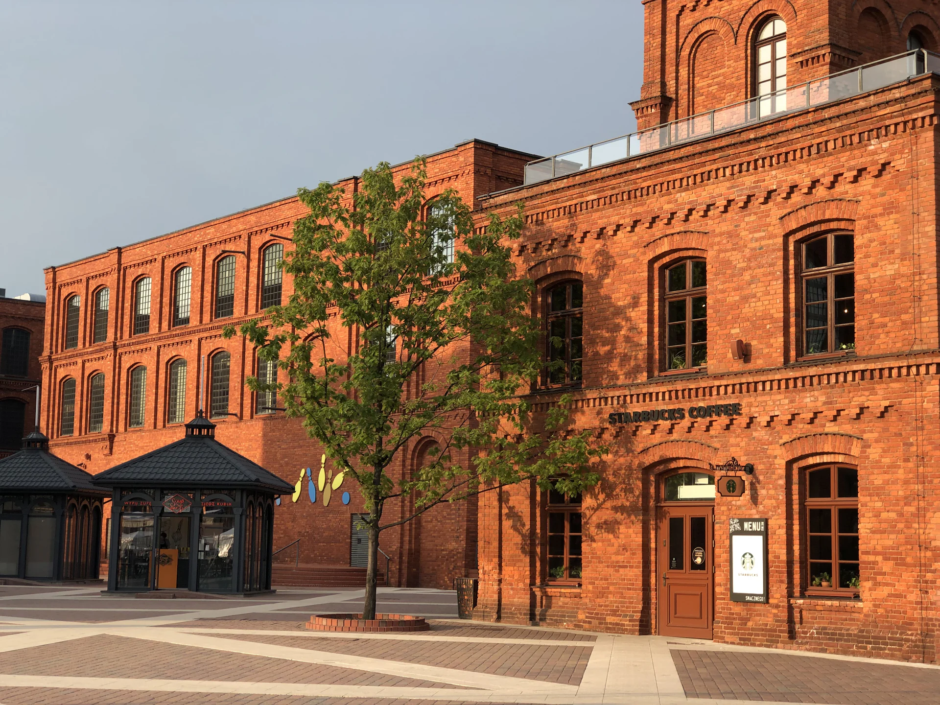 Ceglany budynek dawnej fabryki otoczony zielonymi drzewami