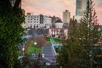 Urban setting with a blend of residential and commercial buildings under a pinkish evening sky. A clock tower stands prominently on a landscaped island amid roads and pathways. Leafy trees frame the scene, and a street with cars and signs adds to the city atmosphere.