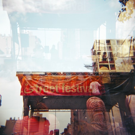 A double-exposure photograph showcasing an outdoor event with a large orange banner reading 'street festival.' In the background, a stage with equipment, possibly a sound system, is visible along with silhouettes of people. Various structures and a clear sky with clouds add to the layered composition.