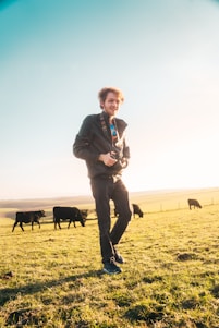A person standing in a sunny field, holding a camera. The scene includes several grazing cows in the background, suggesting a rural setting with open, grassy spaces and clear blue skies.