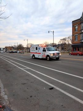 An ambulance with flashing lights is traveling quickly down a multi-lane road, with some traffic visible in the distance. The landscape includes buildings, bare trees, and a cloudy sky, suggesting an urban setting.