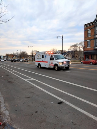 An ambulance with flashing lights is traveling quickly down a multi-lane road, with some traffic visible in the distance. The landscape includes buildings, bare trees, and a cloudy sky, suggesting an urban setting.