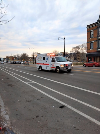 An ambulance with flashing lights is traveling quickly down a multi-lane road, with some traffic visible in the distance. The landscape includes buildings, bare trees, and a cloudy sky, suggesting an urban setting.