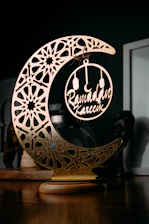 A decorative piece featuring an ornate, crescent moon with intricate geometric patterns and a circular ornament hanging in its center, containing the words 'Ramadan Kareem'. The item is placed on a wooden surface with a dark background, highlighting the gold and metallic elements of the decoration.