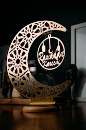 A decorative piece featuring an ornate, crescent moon with intricate geometric patterns and a circular ornament hanging in its center, containing the words 'Ramadan Kareem'. The item is placed on a wooden surface with a dark background, highlighting the gold and metallic elements of the decoration.