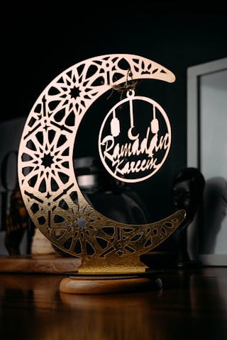 A decorative piece featuring an ornate, crescent moon with intricate geometric patterns and a circular ornament hanging in its center, containing the words 'Ramadan Kareem'. The item is placed on a wooden surface with a dark background, highlighting the gold and metallic elements of the decoration.