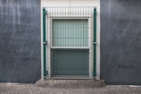 A window is secured with a metal grid situated on a concrete wall. The surrounding area appears to have a textured, rough surface, with subtle streaks indicating possible weather exposure.