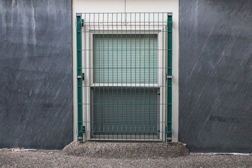 A window is secured with a metal grid situated on a concrete wall. The surrounding area appears to have a textured, rough surface, with subtle streaks indicating possible weather exposure.