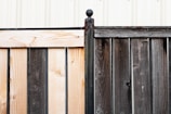 A wooden fence composed of two sections with contrasting colors, one side is light wood while the other is dark. The sections are joined by a rusted metal post capped with a spherical finial. In the background, a white corrugated metal wall is visible, adding a structured, industrial feel.