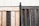 A wooden fence composed of two sections with contrasting colors, one side is light wood while the other is dark. The sections are joined by a rusted metal post capped with a spherical finial. In the background, a white corrugated metal wall is visible, adding a structured, industrial feel.