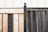A wooden fence composed of two sections with contrasting colors, one side is light wood while the other is dark. The sections are joined by a rusted metal post capped with a spherical finial. In the background, a white corrugated metal wall is visible, adding a structured, industrial feel.