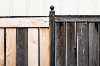 A wooden fence composed of two sections with contrasting colors, one side is light wood while the other is dark. The sections are joined by a rusted metal post capped with a spherical finial. In the background, a white corrugated metal wall is visible, adding a structured, industrial feel.