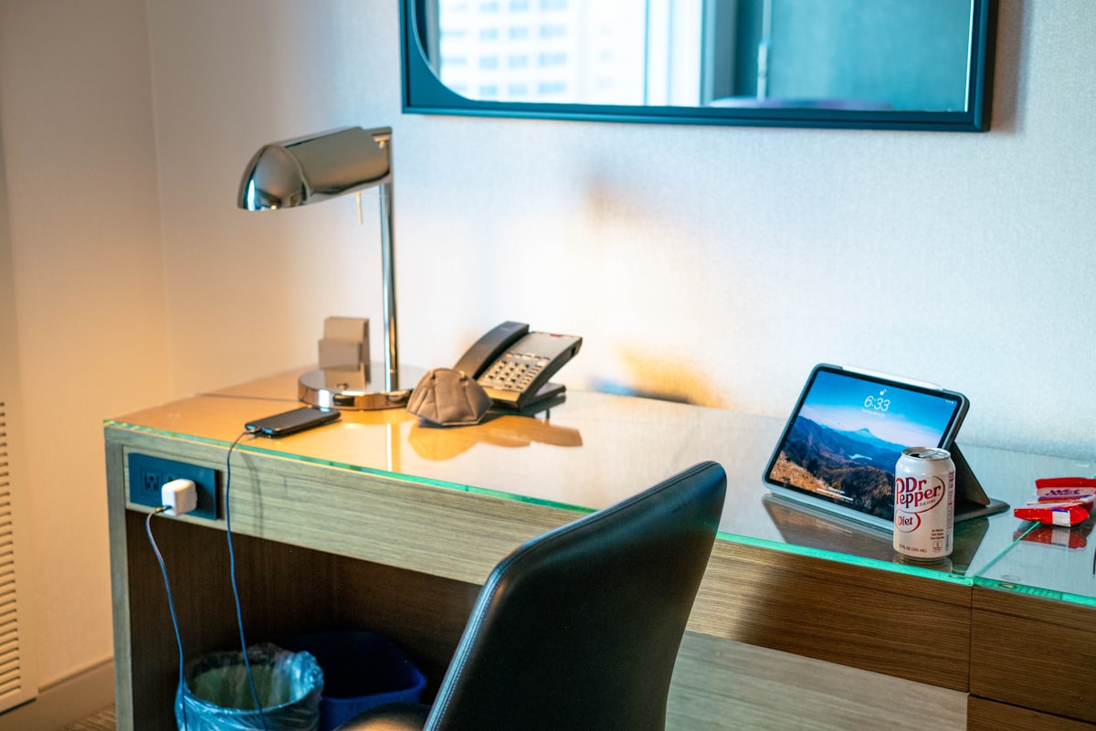A hotel room desk with an adjustable reading lamp, a telephone, a tablet on a stand, a black office chair and a window looking out over the city