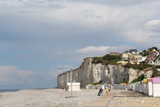 A quiet beach scene featuring white chalk cliffs on the right with several houses perched above. A line of colorful beach cabins with pastel roofs stands on the sandy shore. The sky is partly cloudy with patches of blue visible. Few people can be seen walking or sitting along the boardwalk that stretches parallel to the cabins.