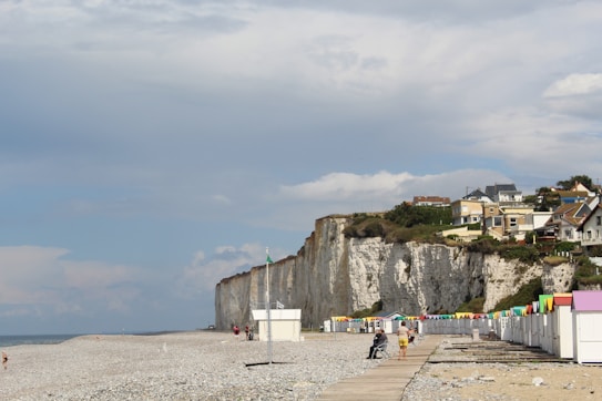 A quiet beach scene featuring white chalk cliffs on the right with several houses perched above. A line of colorful beach cabins with pastel roofs stands on the sandy shore. The sky is partly cloudy with patches of blue visible. Few people can be seen walking or sitting along the boardwalk that stretches parallel to the cabins.