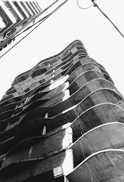 An upward perspective of a multi-story building with a unique undulating architectural design, featuring exposed concrete and brickwork. Power lines cross the upper part of the frame, adding an urban element.