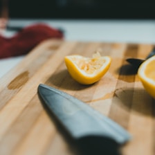 A wooden cutting board with a sharp knife lying next to a sliced lemon. The lemon is cut in half, revealing its juicy interior. The cutting board has a few juice stains, and the scene is softly lit, creating a calm and focused atmosphere.
