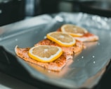 Three slices of salmon are arranged on a sheet of aluminum foil, each topped with a slice of lemon. The background is slightly out of focus, highlighting the fish in the foreground.