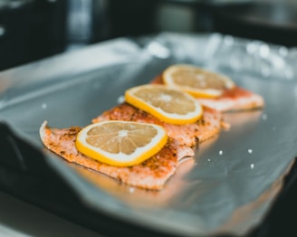 Three slices of salmon are arranged on a sheet of aluminum foil, each topped with a slice of lemon. The background is slightly out of focus, highlighting the fish in the foreground.