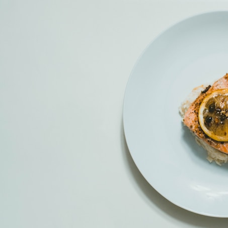 A minimalistic arrangement of a salmon dish on a white plate. The salmon is garnished with a slice of lemon and a few capers, placed on a bed of rice. The setup is simple and elegant, against a light background.