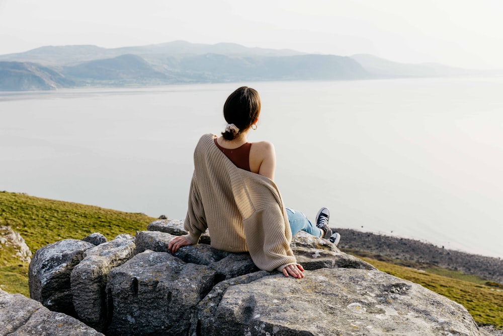 Mujer con camisa marrón de manga larga sentada en la roca cerca del cuerpo de agua durante el día
