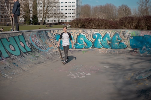 A young person is walking inside a graffiti-covered skate park bowl holding a skateboard. Another person stands on the edge watching. The background includes trees, a building, and a grassy area with people sitting.