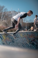 A young person is skateboarding on a ramp with colorful graffiti in an outdoor skate park. Another person is standing nearby with a skateboard, watching the action. The scene is set against a clear blue sky and bare trees, suggesting a chilly but sunny day.