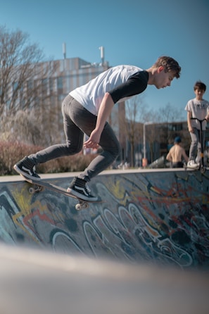 A young person is skateboarding on a ramp with colorful graffiti in an outdoor skate park. Another person is standing nearby with a skateboard, watching the action. The scene is set against a clear blue sky and bare trees, suggesting a chilly but sunny day.