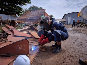 A welder is working on a piece of metal on a construction site, wearing protective gear including a welding helmet and gloves. There are other workers in the background, some wearing hard hats, amidst various metal structures and industrial equipment. The ground is muddy, and a large rusted metal structure is visible in the background.