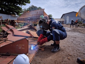A welder is working on a piece of metal on a construction site, wearing protective gear including a welding helmet and gloves. There are other workers in the background, some wearing hard hats, amidst various metal structures and industrial equipment. The ground is muddy, and a large rusted metal structure is visible in the background.