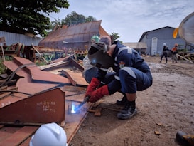 A welder is working on a piece of metal on a construction site, wearing protective gear including a welding helmet and gloves. There are other workers in the background, some wearing hard hats, amidst various metal structures and industrial equipment. The ground is muddy, and a large rusted metal structure is visible in the background.