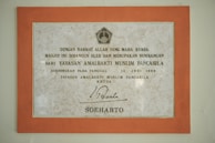 A framed plaque with a beige marble pattern background and a bold orange border featuring Indonesian text. The plaque is a commemoration from Yayasan Amalbakti Muslim Pancasila, dated June 14, 1996, and includes a signature at the bottom.