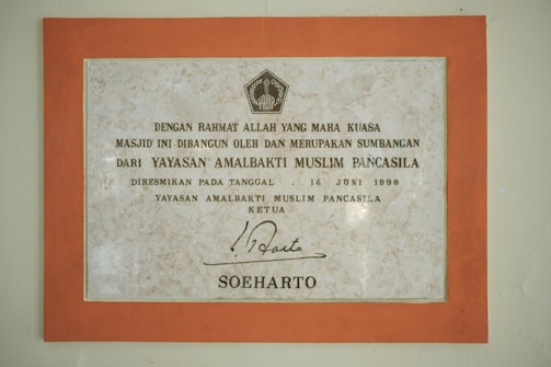 A framed plaque with a beige marble pattern background and a bold orange border featuring Indonesian text. The plaque is a commemoration from Yayasan Amalbakti Muslim Pancasila, dated June 14, 1996, and includes a signature at the bottom.