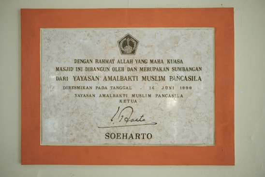 A framed plaque with a beige marble pattern background and a bold orange border featuring Indonesian text. The plaque is a commemoration from Yayasan Amalbakti Muslim Pancasila, dated June 14, 1996, and includes a signature at the bottom.