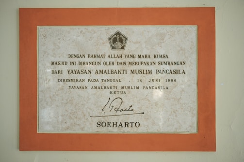 A framed plaque with a beige marble pattern background and a bold orange border featuring Indonesian text. The plaque is a commemoration from Yayasan Amalbakti Muslim Pancasila, dated June 14, 1996, and includes a signature at the bottom.