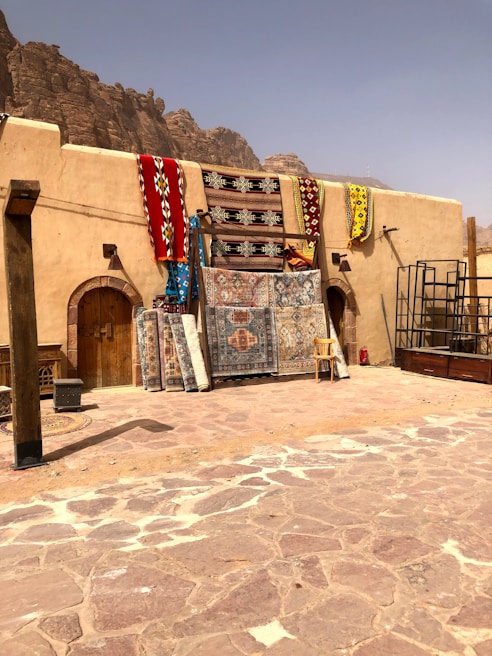 Several colorful rugs and carpets are displayed hanging against a rustic, beige wall. The area is surrounded by a rocky, desert landscape, suggesting a market or shop in a warm, arid region. A chair and some metal frames are also visible, with large rocky hills in the background under a hazy sky.
