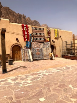 Several colorful rugs and carpets are displayed hanging against a rustic, beige wall. The area is surrounded by a rocky, desert landscape, suggesting a market or shop in a warm, arid region. A chair and some metal frames are also visible, with large rocky hills in the background under a hazy sky.