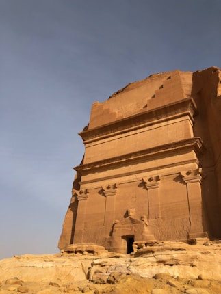 An ancient rock-cut structure with elaborate carvings is situated in a desert landscape. The building appears to be a tomb or temple carved into a large rock formation, displaying intricate details typical of ancient architecture.