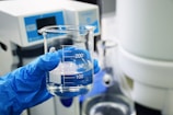 A gloved hand holds a clear glass beaker with water, against a laboratory background with scientific equipment.