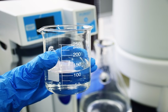 A gloved hand holds a clear glass beaker with water, against a laboratory background with scientific equipment.