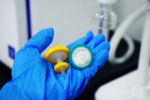A gloved hand holds two medical filter devices, one yellow and one green. The background features lab equipment and a neutral setting.