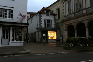 A quaint street scene featuring a mix of historic and modern architecture. A small shop with a brightly lit display window showcases colorful products. On the left, a building displays a British flag. The surroundings include cobblestone pavement and decorative greenery, with a stone structure in front of a columned building.