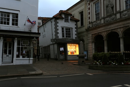 A quaint street scene featuring a mix of historic and modern architecture. A small shop with a brightly lit display window showcases colorful products. On the left, a building displays a British flag. The surroundings include cobblestone pavement and decorative greenery, with a stone structure in front of a columned building.