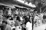 People participate in a bustling outdoor market with numerous stalls selling various fruits. The scene is busy with many individuals browsing and purchasing goods. Bunches of bananas and other tropical fruits hang prominently at the vendor stands.